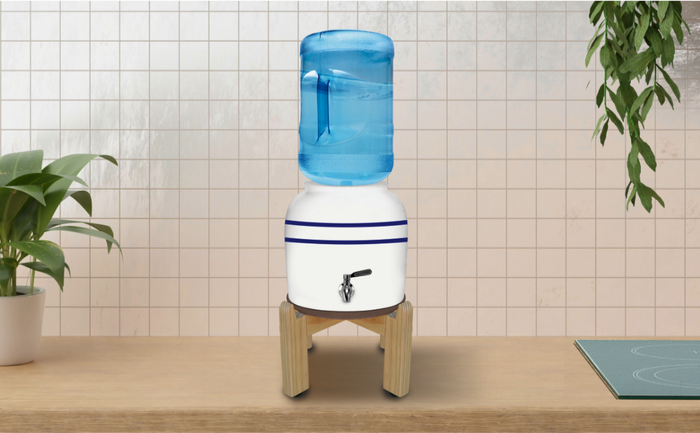 White ceramic water dispenser with blue stripes on a wooden stand, topped with a large blue water jug, placed on a wooden countertop with plants on both sides against a tiled wall.