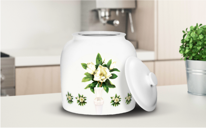 White ceramic water dispenser with floral design featuring white flowers and green leaves, accompanied by a matching lid resting on the side, placed on a kitchen counter with a potted plant in the background.