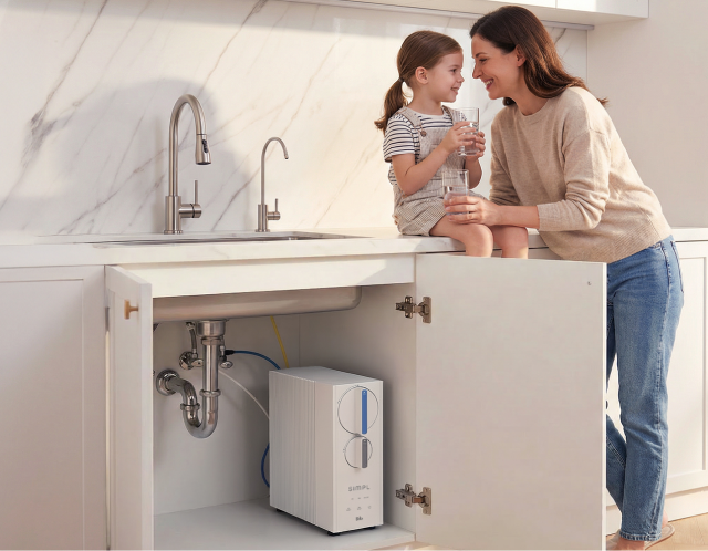 Bright modern kitchen with a mother and young daughter smiling and holding glasses of water by the sink, with a Brio SIMPL RO undersink water filtration system visible inside an open cabinet.