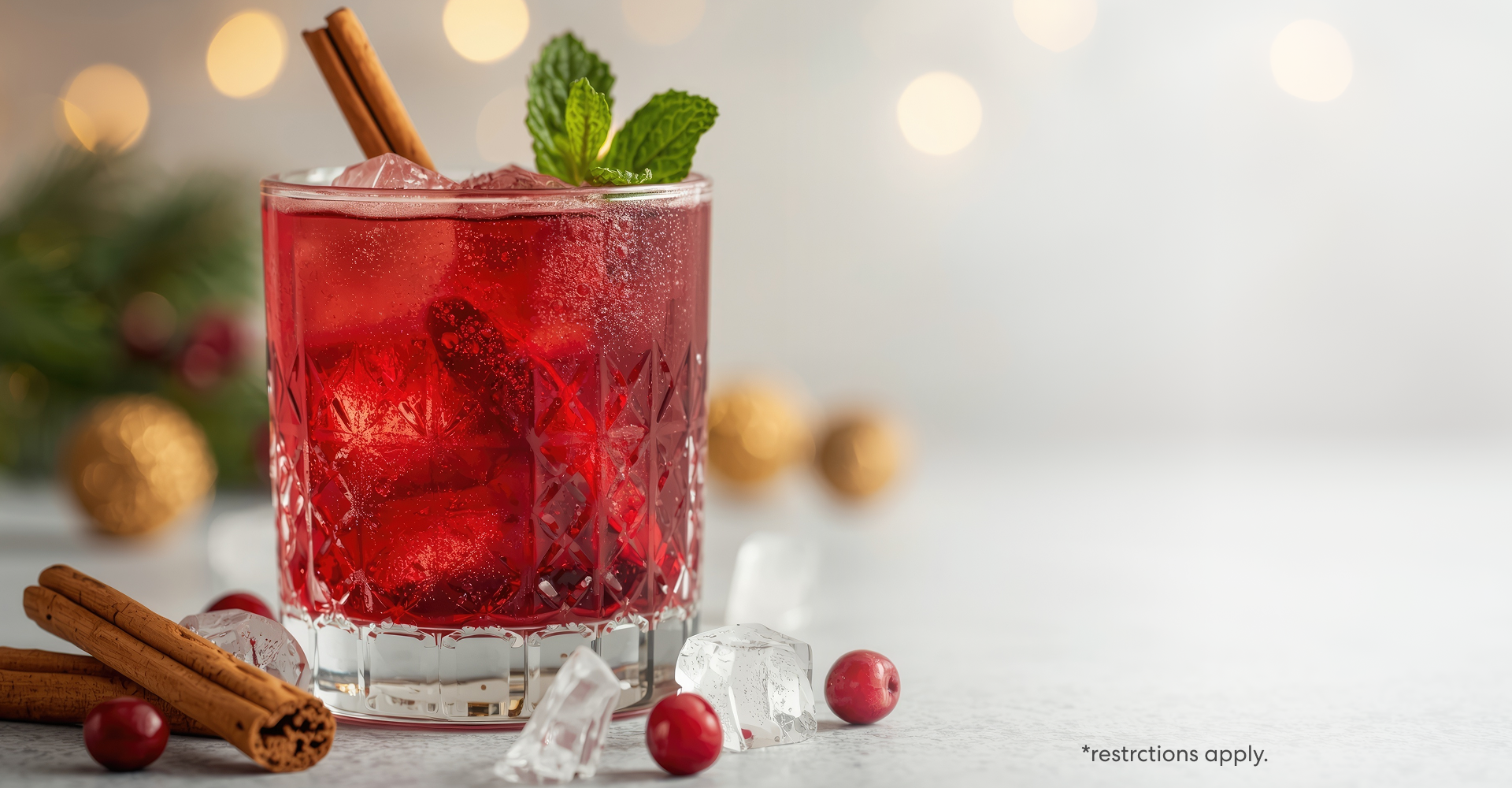 Festive holiday drink in a crystal glass filled with red cranberry spritzer, ice cubes, cinnamon sticks, and mint garnish, surrounded by cranberries and holiday décor on a bright winter background. Restrictions apply.
