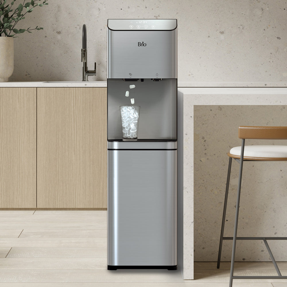 A stainless steel Brio Moderna + Ice water dispenser drops bullet-shaped ice cubes into a clear glass in a modern kitchen with light wood cabinets, a stone countertop and island, and a bar stool beside it.