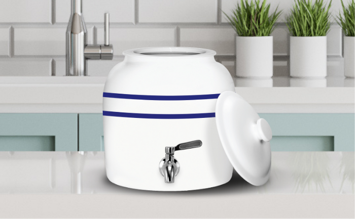 White ceramic water dispenser with two blue stripes and a silver spigot, placed on a kitchen counter with its lid resting on the side, in front of a sink and potted plants.