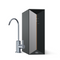 A modern black Brio Tankless RO Under Sink Filtration System 600GPD - PRISM Series with a sleek rectangular design stands next to a curved silver faucet. This advanced unit features reverse osmosis filtration and displays indicator lights labeled PCB, RO, and Reset on the front panel, with the brand name Brio positioned at the bottom.