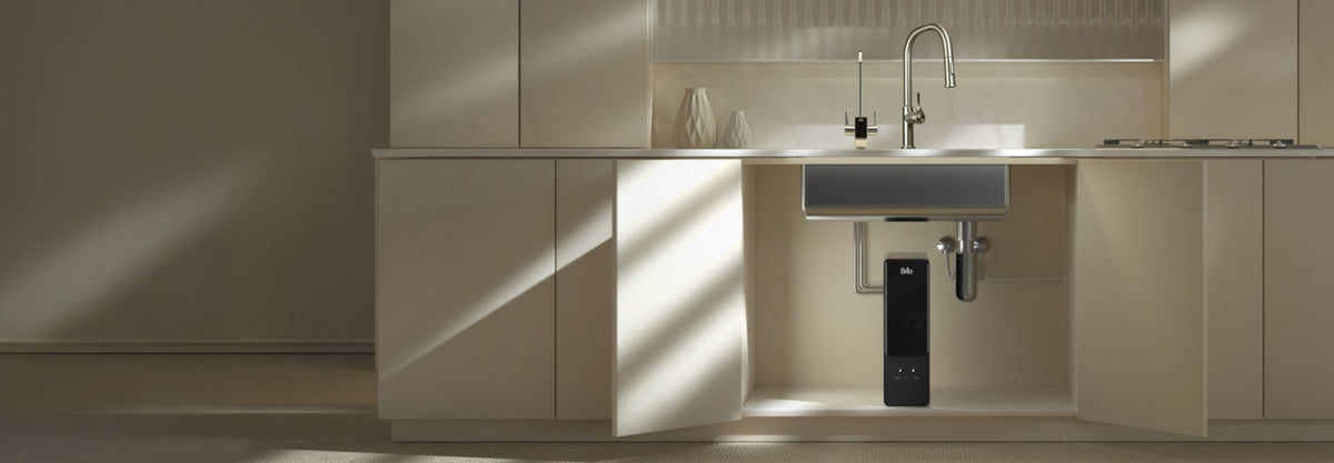 A black Brio Aquus under-sink water filtration system sits inside an open kitchen cabinet beneath a stainless steel sink. Above the counter, its smart faucet with a digital display stands beside the main kitchen faucet, in a warm, sunlit kitchen with cream cabinets.