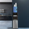 A Top Load Water Cooler - 420 Series, equipped with a blue water jug and a stack of white cups on top, featuring stainless steel reservoirs, is placed against a dark wall in a modern kitchen with black cabinets and open shelving in the background.