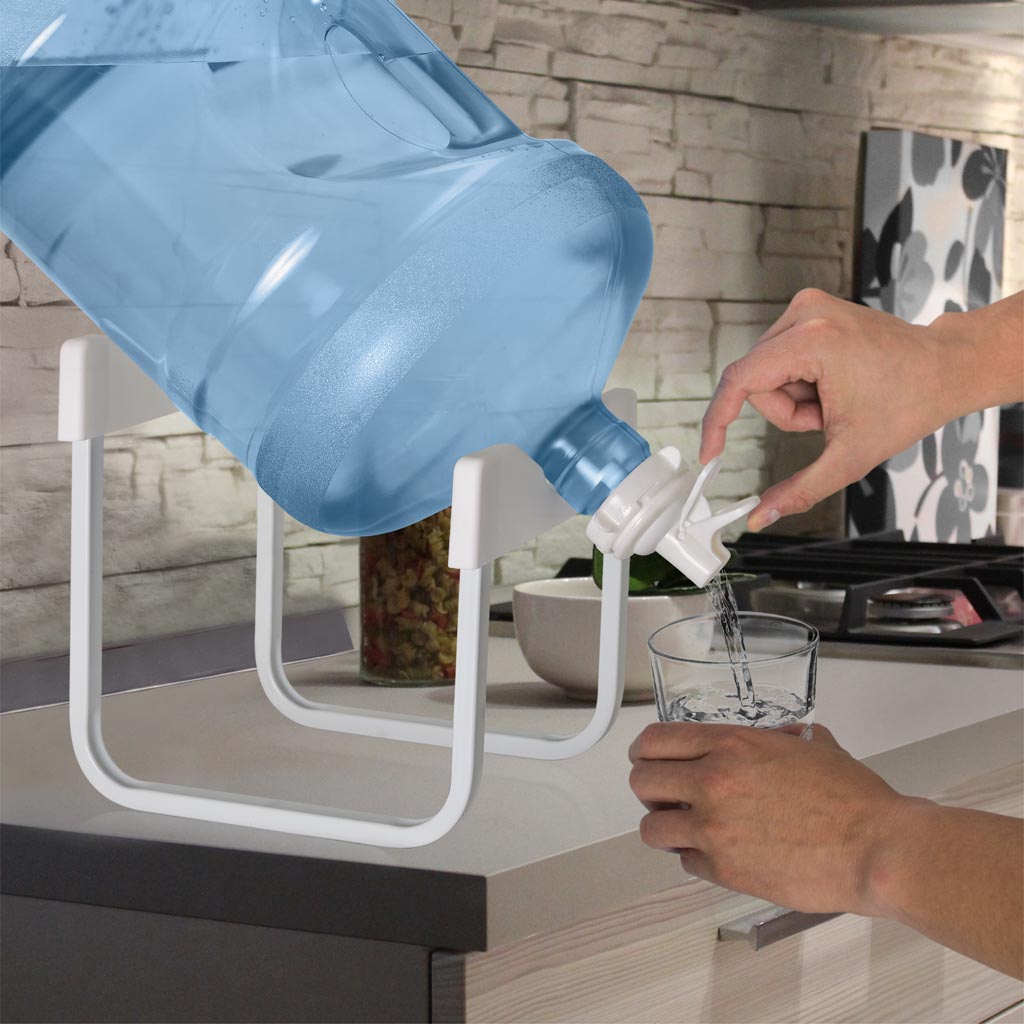 A person fills a clear glass with water from a 5-gallon blue bottle on the Stainless Steel Single Water Bottle Stand - White, placed next to a stainless steel sink on the kitchen counter.