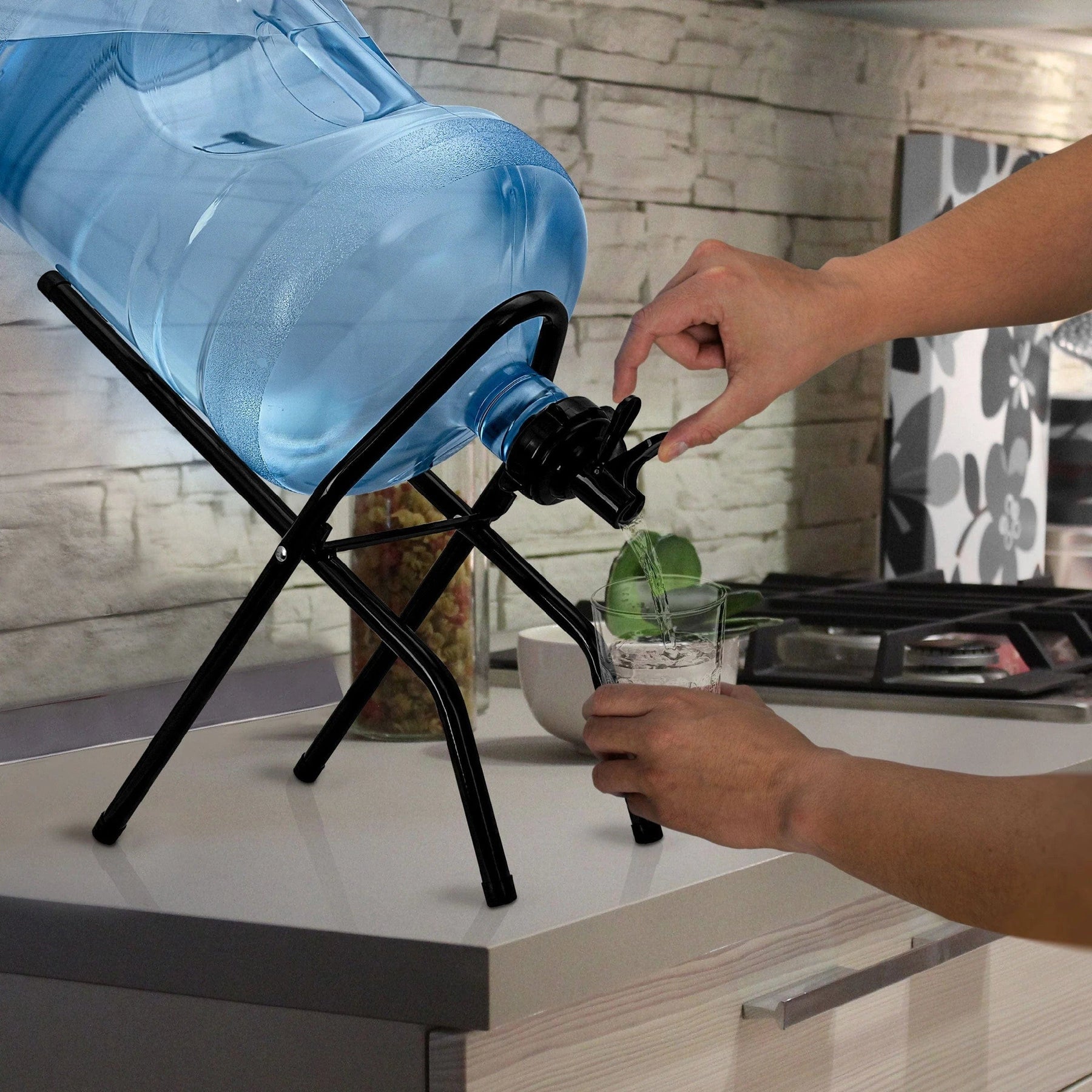 Using the Brio Metal Foldable Bottle Stand – Black, a person fills a glass with water from a large blue 5-gallon bottle in a modern kitchen.