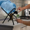A person fills a glass of water from a dispenser placed on the Metal Foldable Bottle Stand - Black in a modern kitchen.