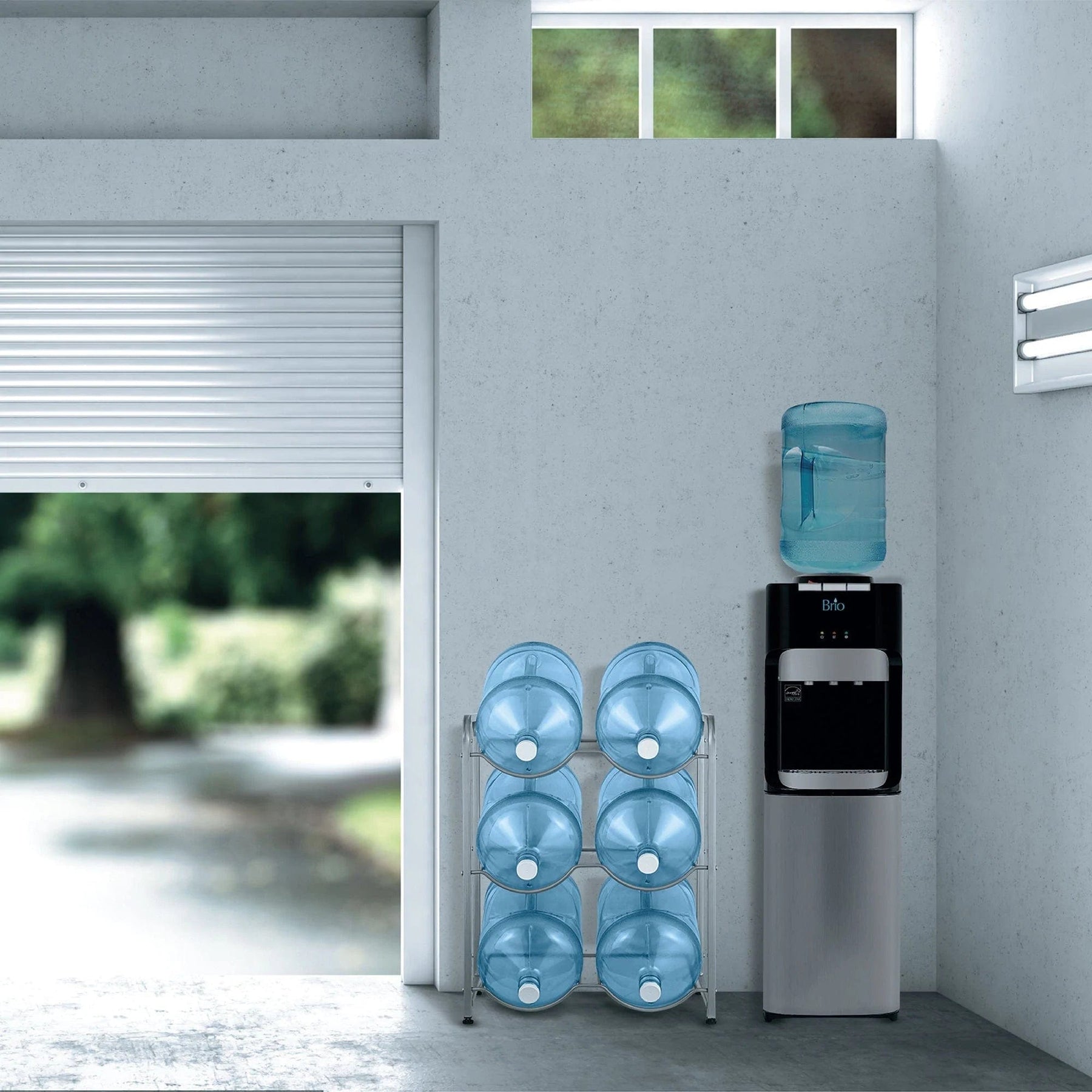 A modern room features the Brio Double Column 6-Bottle Storage Rack in gray with a blue water jug, efficiently storing six large blue bottles. A window and open garage door reveal greenery outside.