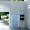 A modern room features a stainless steel water dispenser with a blue jug, a Double Column 8-Bottle Storage Rack - White stacked with eight empty blue jugs, and a large window letting in natural light.