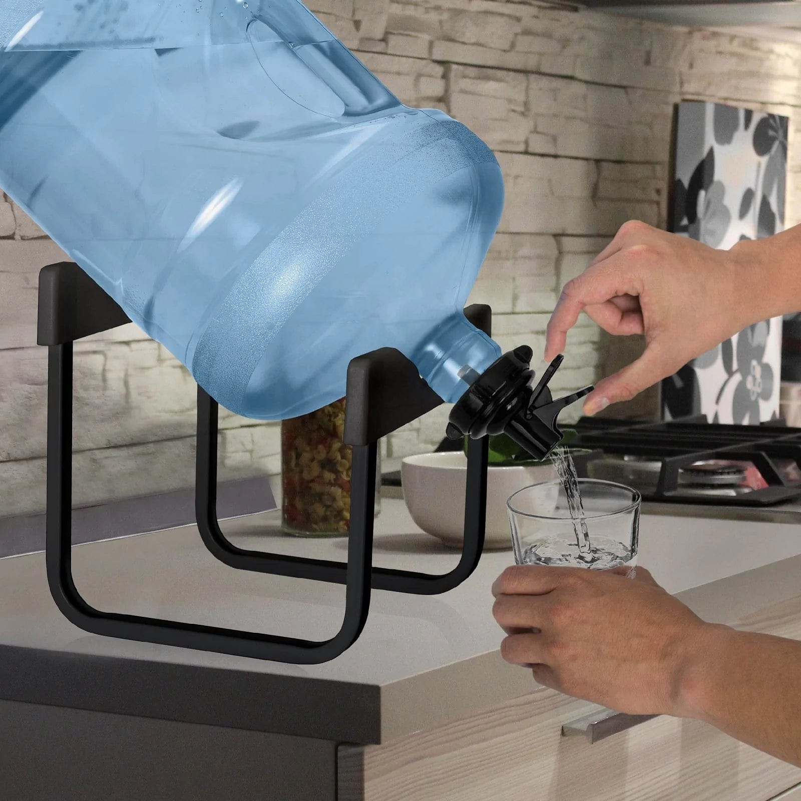 A person uses both hands to dispense water from a Brio Stainless Steel Single Water Bottle Stand - Black, holding a large blue 3 or 5-gallon bottle, into a glass on the kitchen counter with a stove and food jar in the background.