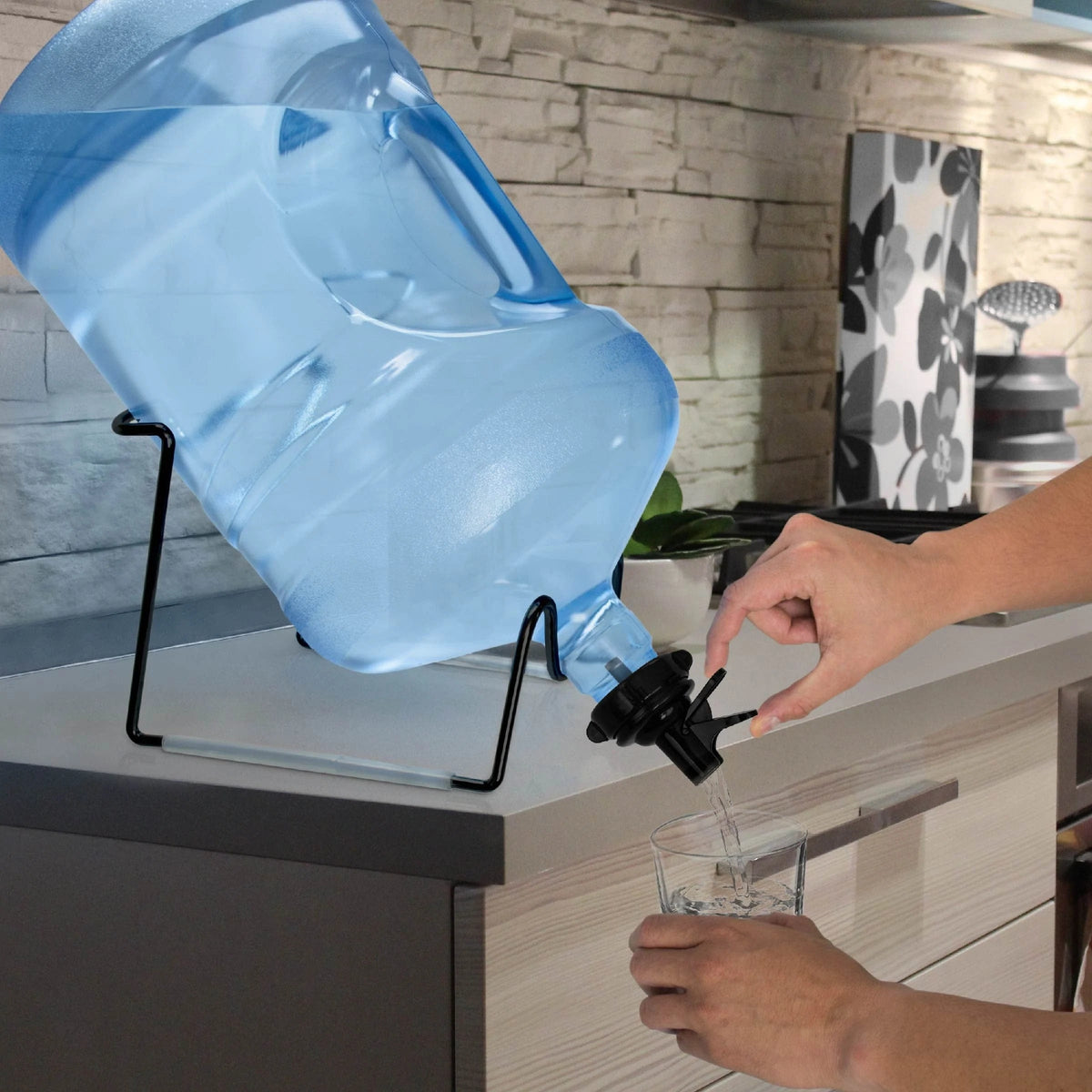 A person fills a glass with water from a large blue jug placed on the Metal-Plated Water Bottle Stand - Black, set on the kitchen counter against a stone backsplash with various kitchen items in the background.